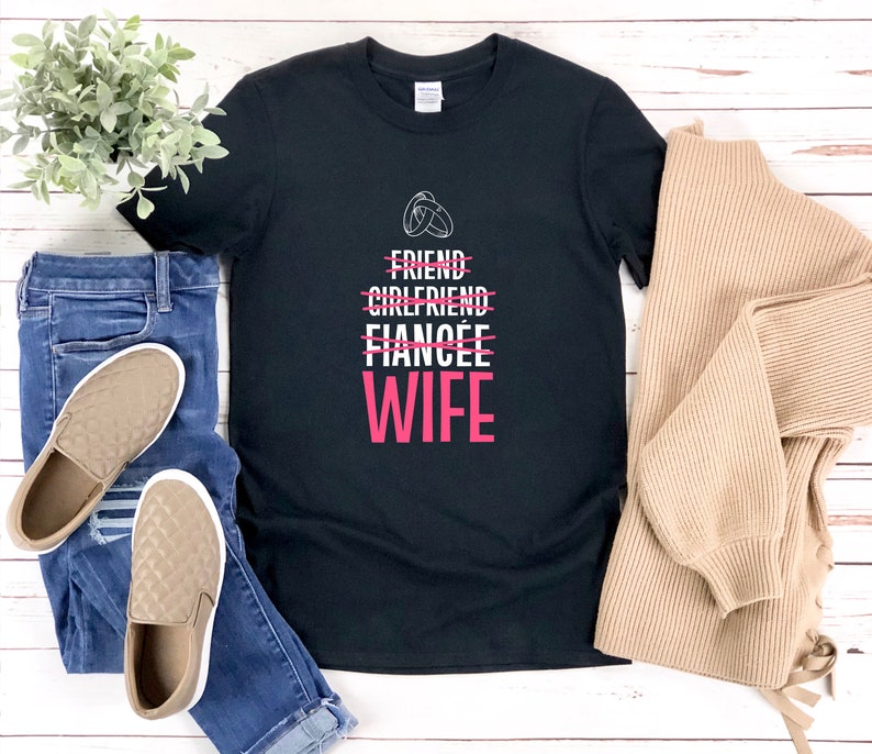 Puede incluir: Camiseta negra con texto blanco que dice "Friend, Girlfriend, Fianc&eacute;e" con una l&iacute;nea a trav&eacute;s de cada palabra. La palabra "Wife" est&aacute; en rosa debajo de las otras palabras. La camiseta est&aacute; sobre una superficie de madera blanca con un par de zapatos acolchados marrones y unos vaqueros azules.