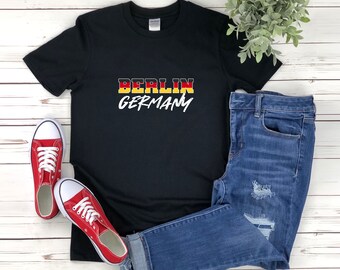 Puede incluir: Camiseta negra con la palabra "BERLIN" en rojo, amarillo y negro, y la palabra "GERMANY" en escritura blanca. La camiseta se combina con vaqueros azules y zapatillas rojas con cordones blancos.