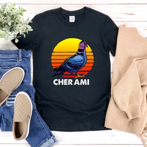 May include: Black t-shirt featuring a colorful illustration of a pigeon against a sunset backdrop. The design includes the text "CHER AMI" in white. The shirt is paired with blue jeans and tan slip-on shoes.