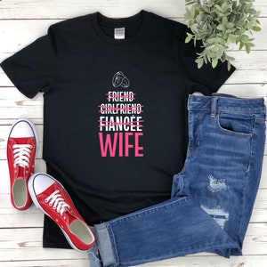 Puede incluir: Camiseta negra con texto blanco que dice "Friend, Girlfriend, Fianc&eacute;e, Wife" con una l&iacute;nea que tacha las tres primeras palabras y la palabra "Wife" en rosa. La camiseta est&aacute; sobre una superficie de madera blanca con un par de zapatillas rojas y unos vaqueros azules.