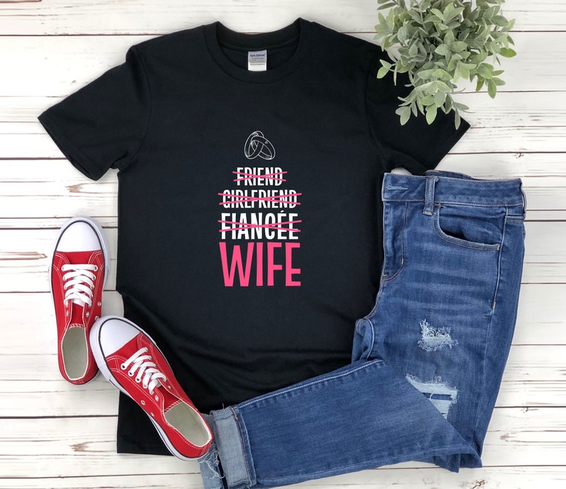 Puede incluir: Camiseta negra con texto blanco que dice "Friend, Girlfriend, Fianc&eacute;e, Wife" con una l&iacute;nea que tacha las tres primeras palabras y la palabra "Wife" en rosa. La camiseta est&aacute; sobre una superficie de madera blanca con un par de zapatillas rojas y unos vaqueros azules.