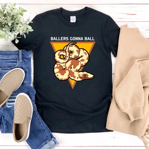Op de afbeelding: Zwart T-shirt met een gele driehoek en een bruine en witte slang erin. De tekst "Ballers Gonna Ball" staat boven de driehoek.