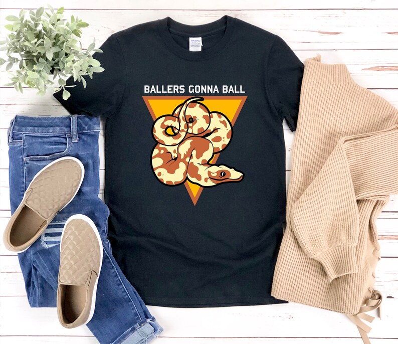 Op de afbeelding: Zwart T-shirt met een gele driehoek en een bruine en witte slang erin. De tekst "Ballers Gonna Ball" staat boven de driehoek.