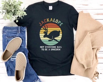 May include: Black t-shirt featuring a jackalope graphic within a circular design with a sunset background. The text "JACKALOPE" arches above the image, and below it reads "NOT EVERYONE HAS TO BE A UNICORN". The shirt is paired with blue jeans and tan slip-on shoes.