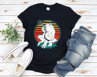 Könnte beinhalten: Ein schwarzes T-Shirt mit einem Retro-Grafikdruck eines Frosches, der Banjo spielt. Der Frosch ist weiß und sitzt auf einem Pilz mit einem blauen und grünen Gittermuster dahinter. Der Hintergrund ist ein Sonnenuntergang mit orangefarbenen, roten und gelben Streifen.