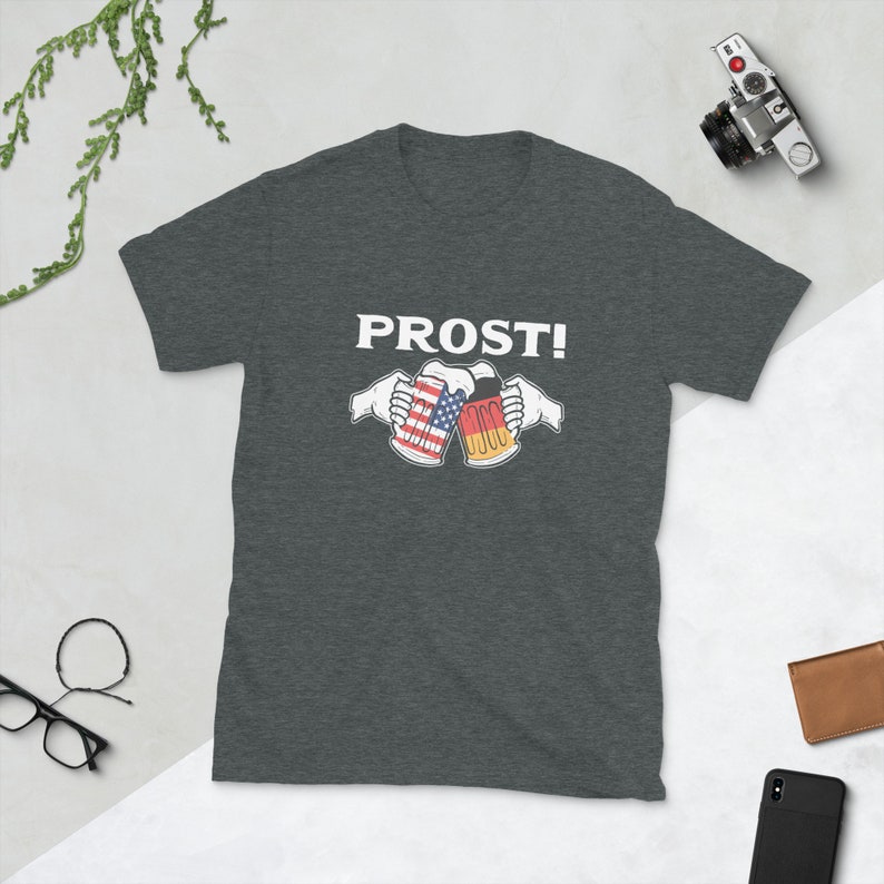 Pode incluir: Uma camiseta cinza escura com o texto "PROST!" em branco. Duas canecas de cerveja brindando, uma com a bandeira americana e a outra com a bandeira alem&atilde;.
