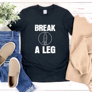 Puede incluir: Camiseta negra con texto blanco que dice "Break a Leg" y un gráfico de un hueso de pierna roto en un círculo.