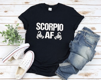 May include: Black t-shirt with the text "SCORPIO AF" in white, along with two white scorpion graphics. The shirt is paired with blue jeans and white canvas sneakers with red and blue accents. The items are arranged on a light wood-grain surface.