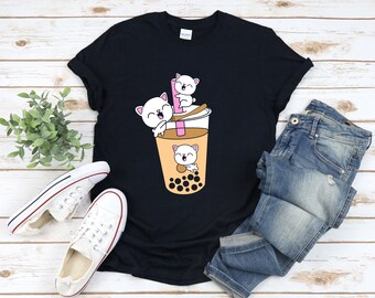 May include: Black t-shirt featuring a graphic of three white cats in a bubble tea cup. The cats are smiling and have pink noses. The bubble tea cup is filled with brown liquid and boba pearls.