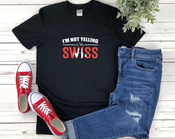 Può includere: Maglietta nera con testo bianco che recita "Non sto urlando - Sono svizzero" in lettere rosse. La maglietta è stesa su una superficie di legno bianco con un paio di scarpe da ginnastica rosse e jeans blu.