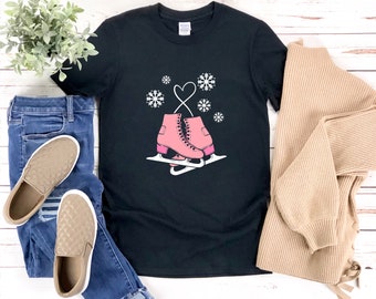 以下が含まれることがあります： ピンクのスケートのグラフィックと雪の結晶が描かれた黒いTシャツ。グラフィックは、ハートの形で結ばれた2つのスケートを示しています。