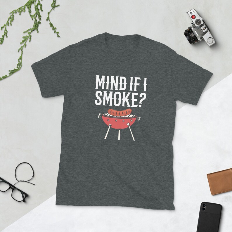 May include: A dark heather grey t-shirt with the text "Mind if I Smoke?" in white. A cartoon image of a red grill with a sausage on it is below the text.