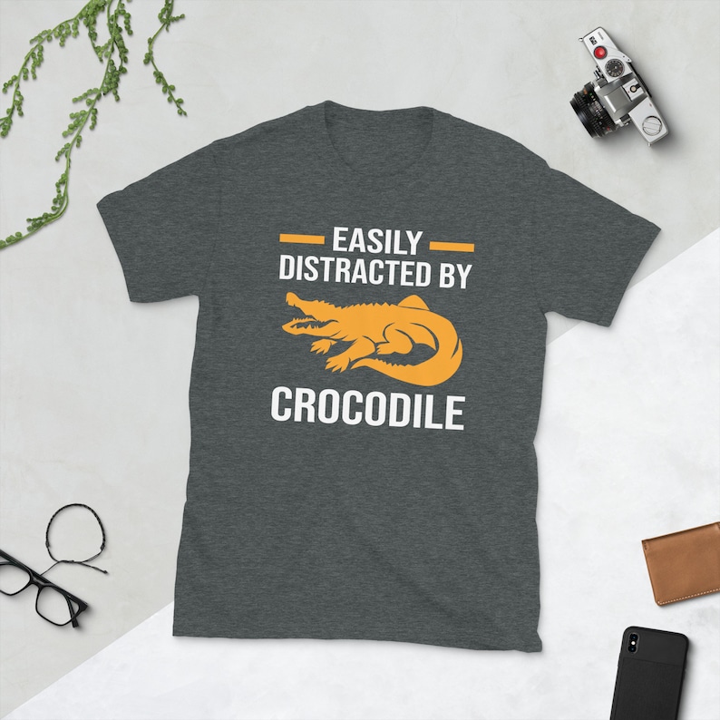 Op de afbeelding: Donkergrijs t-shirt met de tekst "EASILY DISTRACTED BY CROCODILE" in het wit en een oranje alligatorafbeelding. Het shirt is gemaakt van een zacht materiaal en is geschikt voor vrijetijdskleding.
