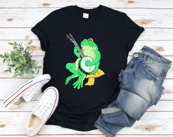 Könnte beinhalten: Schwarzes T-Shirt mit einer Cartoon-Fröschin, die Banjo spielt. Der Frosch ist grün mit gelben Akzenten und sitzt auf einem Pilz. Blaue Jeans und weiße Turnschuhe sind ebenfalls abgebildet.