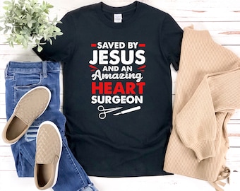 May include: Black t-shirt with white text that reads "Saved by Jesus and an Amazing Heart Surgeon". The text is in a distressed font. There are two white medical tools below the text.