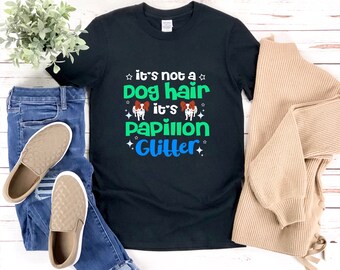 Könnte beinhalten: Schwarzes T-Shirt mit dem Text "it's not a dog hair it's Papillon Glitter" in Grün und Blau. Das Shirt wird mit blauen Jeans, beigen Slip-On-Schuhen und einem beigen Pullover präsentiert.