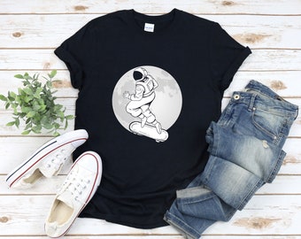 Puede incluir: Una camiseta negra con un gráfico blanco de un astronauta patinando en la luna. El astronauta lleva un casco y un traje espacial. La luna es un círculo con un contorno blanco.