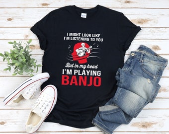 Könnte beinhalten: Ein schwarzes T-Shirt mit weißem Text, der lautet: "Ich sehe vielleicht aus, als würde ich zuhören, aber in meinem Kopf spiele ich Banjo". Über dem Text befindet sich eine Cartoon-Abbildung einer Person, die Banjo spielt.