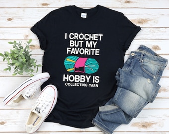May include: Black t-shirt with white text that reads "I crochet but my favorite hobby is collecting yarn". A colorful ball of yarn with knitting needles is pictured on the shirt.