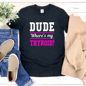 May include: Black t-shirt with the text "DUDE Where's my THYROID?" in white and pink. The shirt is displayed with blue jeans, tan slip-on shoes, and a tan sweater. A small green plant is also in the image.