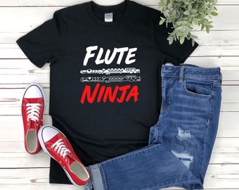 Może przedstawiać: Czarny T-shirt z białym napisem "FLUTE NINJA" i grafiką fletu. Koszulka leży na białym drewnianym tle z parą czerwonych butów sportowych i niebieskimi dżinsami.