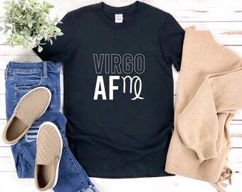 以下が含まれることがあります： 「VIRGO AF」の白文字が入った黒いTシャツ。青いジーンズ、ベージュのスリッポンシューズ、ベージュのセーターと組み合わせています。デザインには乙女座の星座記号が含まれています。