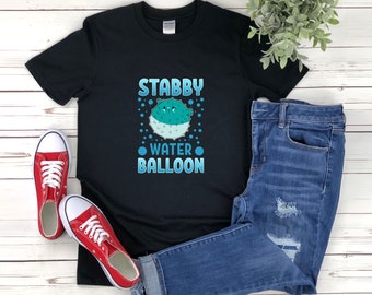 Puede incluir: Camiseta negra con el texto "STABBY WATER BALLOON" y un diseño de pez globo de dibujos animados en color turquesa y blanco. La camiseta se combina con vaqueros azules y zapatillas rojas.