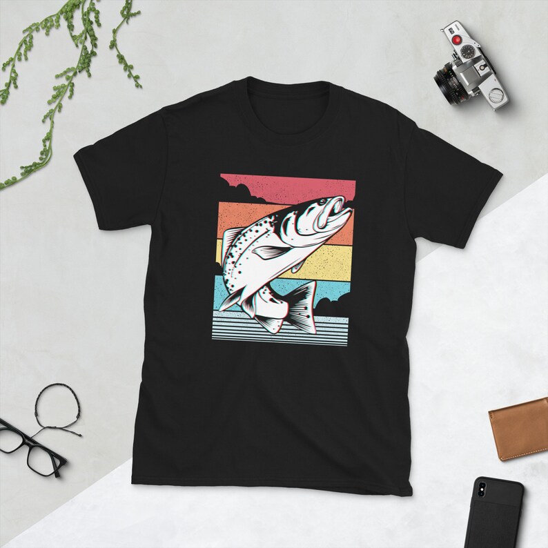 以下が含まれることがあります： 水から飛び出す魚のレトロなグラフィックがプリントされた黒いTシャツ。魚は白で黒いアウトラインがあり、カラフルなストライプの背景に配置されています。
