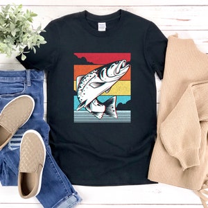 以下が含まれることがあります： 水から飛び出す魚のレトロなグラフィックがプリントされた黒いTシャツ。グラフィックは虹色のカラーリングです。