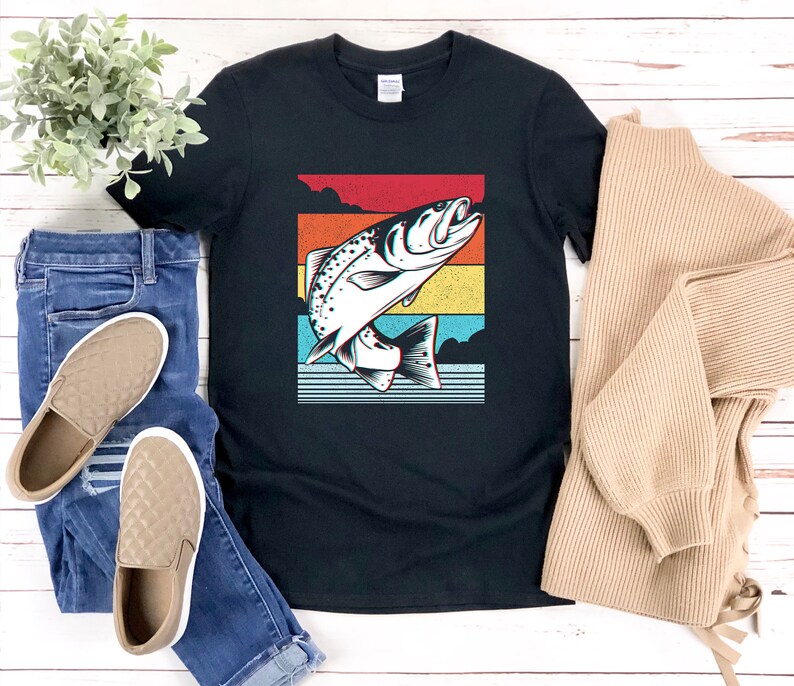 以下が含まれることがあります： 水から飛び出す魚のレトロなグラフィックがプリントされた黒いTシャツ。グラフィックは虹色のカラーリングです。
