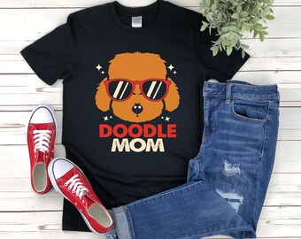 Puede incluir: Camiseta negra con un perro doodle de dibujos animados con gafas de sol rojas y el texto "DOODLE MOM". La imagen también incluye vaqueros azules y zapatillas rojas.