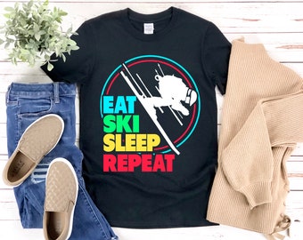 Könnte beinhalten: Schwarzes T-Shirt mit einer bunten Grafik eines Skifahrers in einem Kreis. Der Text "Eat Ski Sleep Repeat" steht unter der Grafik.