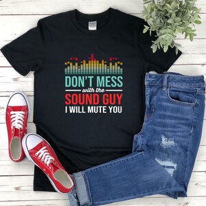 Pode incluir: Camiseta preta com o texto "DON'T MESS with the SOUND GUY I WILL MUTE YOU" e um design de equalizador gráfico. A camiseta é combinada com jeans azuis e tênis vermelhos com solas e cadarços brancos. Um ramo de vegetação está no fundo.