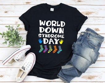 Op de afbeelding: Zwart T-shirt met de tekst "WORLD DOWN SYNDROME DAY" in wit. De tekst wordt omringd door vijf kleurrijke sokken met verschillende patronen.