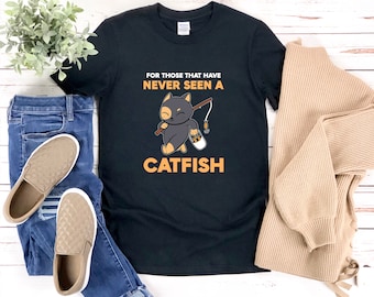 May include: Black t-shirt with a graphic of a cartoon cat holding a fishing rod and a bucket of fish. The text on the shirt reads "For those that have never seen a catfish".