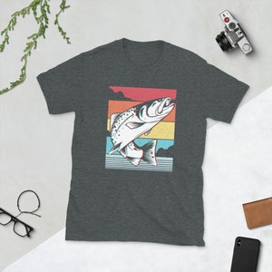 以下が含まれることがあります： 暗いヘザーグレーのTシャツに、水から飛び出す魚のレトロなグラフィックがプリントされています。魚は白で黒いアウトラインがあり、赤、オレンジ、黄色、青、緑のカラフルな背景に配置されています。