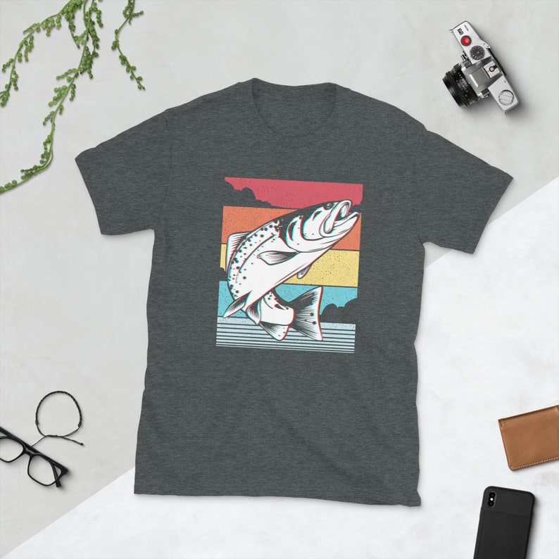 以下が含まれることがあります： 暗いヘザーグレーのTシャツに、水から飛び出す魚のレトロなグラフィックがプリントされています。魚は白で黒いアウトラインがあり、赤、オレンジ、黄色、青、緑のカラフルな背景に配置されています。