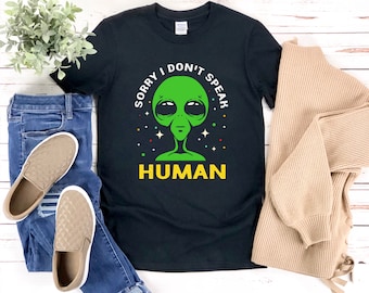 May include: Black t-shirt with a green alien graphic and the text "SORRY I DON'T SPEAK HUMAN" in white and yellow. The alien has large black eyes and a small mouth. The shirt is paired with blue jeans, tan slip-on shoes, and a tan sweater.