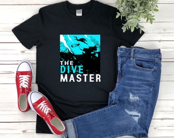 Puede incluir: Camiseta negra con un gráfico de un buceador en agua turquesa. El texto "THE DIVE MASTER" está impreso en blanco debajo del gráfico.