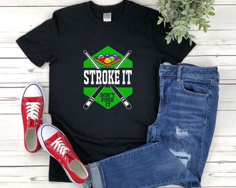 May include: Black t-shirt with a green and white graphic that says "STROKE IT DON'T POKE IT" with pool cues and billiard balls. Red canvas sneakers and blue jeans are also in the image. The shirt is a Gildan brand.