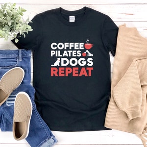 Könnte beinhalten: Schwarzes T-Shirt mit dem Text "COFFEE PILATES DOGS REPEAT" in Rot. Der Text ist mit einer Kaffeetasse, einer Pilates-Pose und einer Hundesilhouette gestaltet.