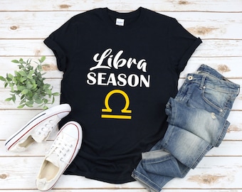 Può includere: Maglietta nera con testo bianco che dice "Libra Season" e un simbolo della Bilancia giallo.