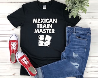 May include: Black t-shirt with white text that reads "Mexican Train Master" and a graphic of four dominoes. The shirt is laying on a white wooden surface with a pair of red sneakers and blue jeans.