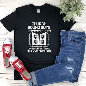 以下が含まれることがあります： 黒いTシャツに白い文字で「Church Sound Guys」と書いてあり、2つのスピーカーのグラフィックがあります。グラフィックの下のテキストには、「Can do a lot of things but they can't put talent in your monitor.」と書いてあります。