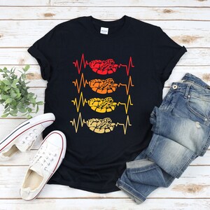 Peut inclure: T-shirt noir avec un motif de serpent en forme de battement de cœur. Le motif représente un serpent dans un motif de battement de cœur avec des couleurs rouge, orange et jaune.