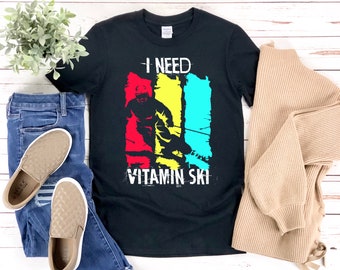 Könnte beinhalten: Schwarzes T-Shirt mit einem Grafikdruck eines Skifahrers in einem farbenfrohen Retro-Design. Der Text "I need Vitamin Ski" ist auf dem Shirt aufgedruckt.