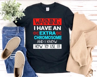 Può includere: Maglietta nera con un cartello di avvertimento rosso e bianco che dice "WARNING I HAVE AN EXTRA CHROMOSOME AND I KNOW HOW TO USE IT!" con un grafico a catena di DNA blu.