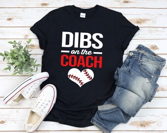 May include: A black t-shirt with white text that says "DIBS on the COACH" in red. A white baseball with red stitching is in the shape of a heart.