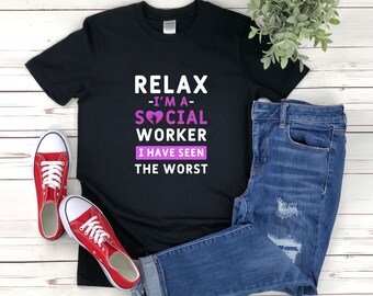 May include: Black t-shirt with the text "Relax - I'm a Social Worker I have seen the worst" in white. A pink broken heart is in the word "Social". The shirt is on a white wooden surface with a pair of red sneakers and blue jeans.