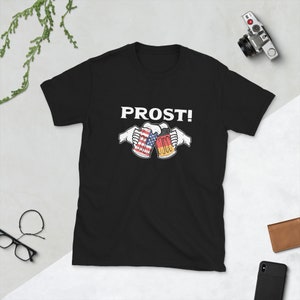 Pode incluir: Camiseta preta com o texto "PROST!" em branco. Duas canecas de cerveja brindando, uma com a bandeira americana e a outra com a bandeira alem&atilde;.